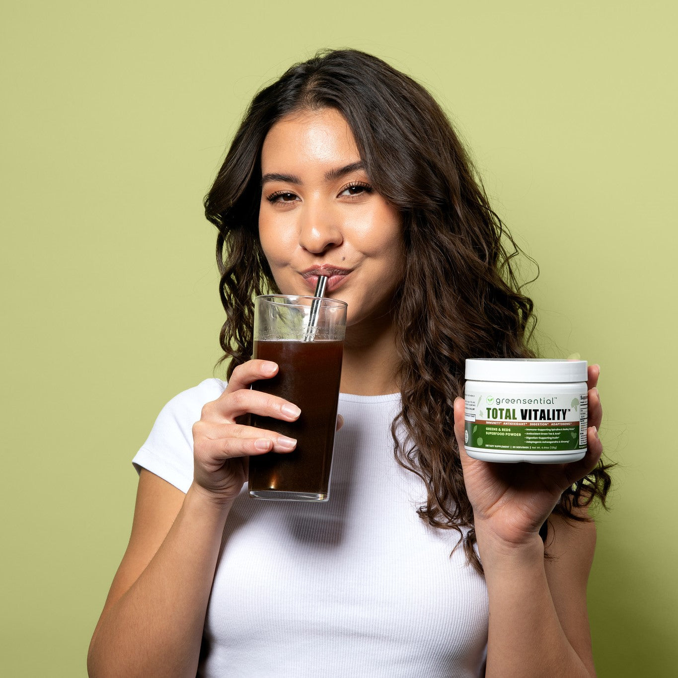 A woman in front of a green backdrop holding the product and drinking from a glass